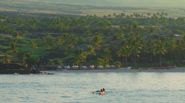 Outrigger canoe with paddlers gliding over calm ocean water at sunrise near palm lined resort beach on Big Island Hawaii with umbrellas colorful kayaks and green lava slopes behind.
