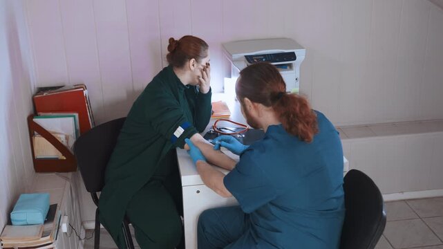 White nurse performing blood test at home desk, redhead clinician stabilizes arm while colleague prepares vials, gloves and needle visible, indoor followup appointment, calm professional care