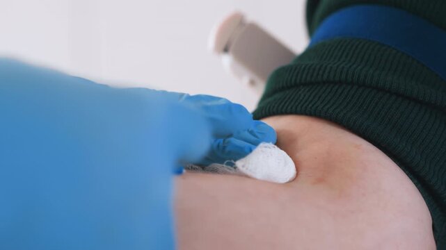 Needle insertion into forearm during blood draw, paramedic inserts needle steadily with gloved hands, small blood drop appears, sterile technique maintained on patient arm over table.