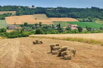 Fototapeta premium Hay bales collecting on a harvested field with a village and green hills in the background under a blue sky