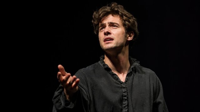 Young male actor in a black shirt performing an emotional dramatic monologue on a dark stage, expressive gesture and intense facial expression under theatrical lighting
