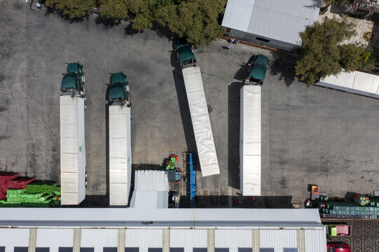 Aerial view of an avocado packing house with semi-trucks parked at loading docks, stacks of plastic crates, and warehouse facilities in Tancitaro, Michoacan, Mexico.