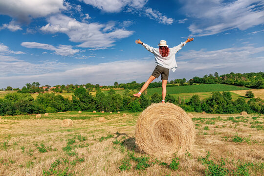 Centered from behind, a red-haired woman balances one-legged on a hay bale with arms wide, green rolling farmland filling the wide frame. Freedom concept.