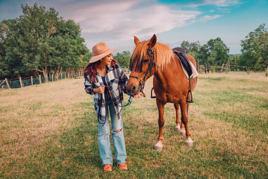 Young woman in a wide-brim hat gently offering her hand to a saddled chestnut horse in a fenced pasture. Country lifestyle concept.