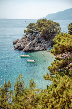 View of the iconic Kamen Brela rock formation surrounded by pine trees, crystal clear turquoise water, and small boats floating near the pebble beach in Brela, Split-Dalmatia County, Croatia.
