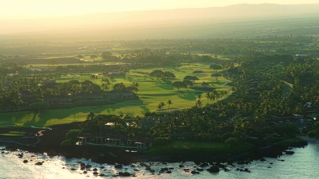 Drone aerial of lush golf fairways and palm trees at sunset on Big Island Hawaii with luxury resort villas lava rock shoreline and glowing Pacific Ocean waves below hazy volcanic slopes.