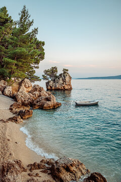 View of the iconic Kamen Brela rock formation surrounded by pine trees, clear turquoise sea, and a small boat near the pebble beach in Brela, Split-Dalmatia County, Croatia.