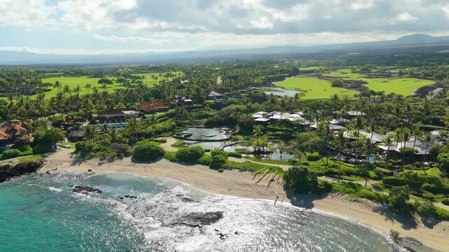 Drone aerial over Big Island Hawaii showing turquoise ocean waves breaking on lava rock reef sandy beach luxury resort villas swaying palm trees and green golf course under volcano slopes.