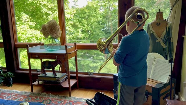 Young boy practicing the trombone indoors near a window