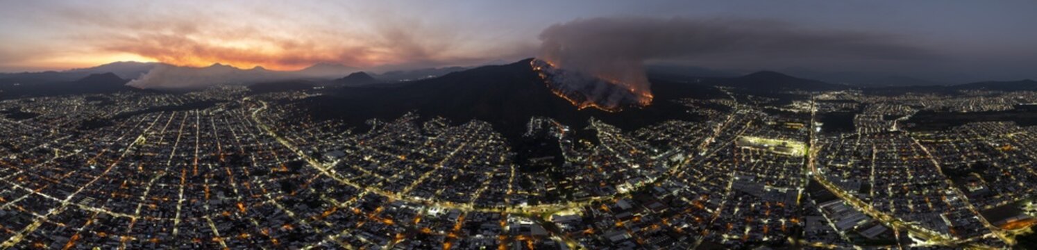 Aerial view of a wildfire burning on the mountainside overlooking the illuminated city grid at dusk, with smoke plumes rising into the twilight sky in Uruapan, Michoacan, Mexico.