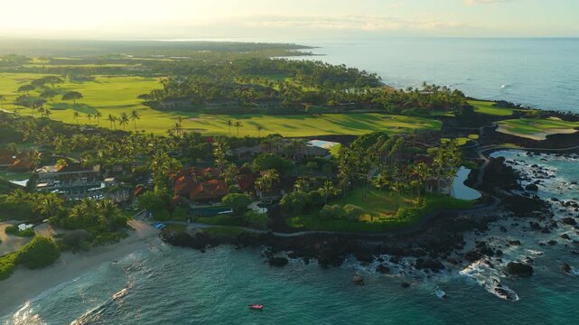 Aerial drone view of luxury oceanfront resort on Big Island Hawaii with palm trees green golf course lava rock coastline white sand beach and turquoise Pacific Ocean waves at golden sunrise.