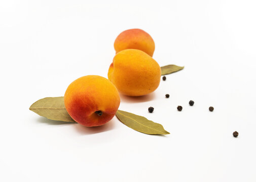 Three apricots with bay leaves and black peppercorns arranged on a white background