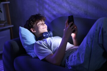 Fototapeta premium Happy teenage boy using mobile phone on sofa at night