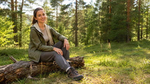 Smiling woman in hiking gear sits on a log in a sunlit pine forest, resting and enjoying nature during a peaceful outdoor hike
