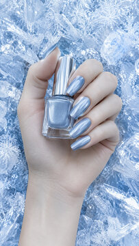 Female hand with metallic silver manicure holding nail polish among ice cubes and frost. Cool winter beauty image for cosmetics, advertising, branding, social media and product presentation.