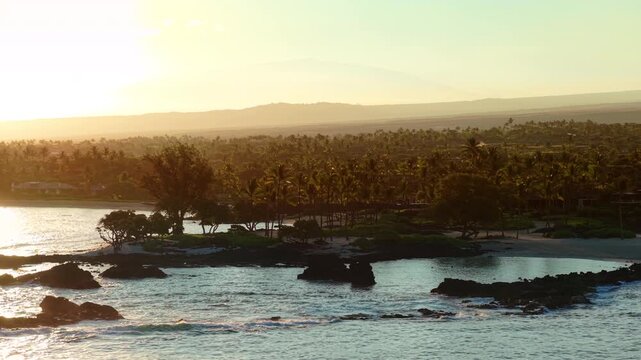 Aerial drone view of golden sunset over rocky lava coastline on Big Island Hawaii with palm groves swaying gently calm turquoise lagoon waves breaking on black rocks and distant volcano.