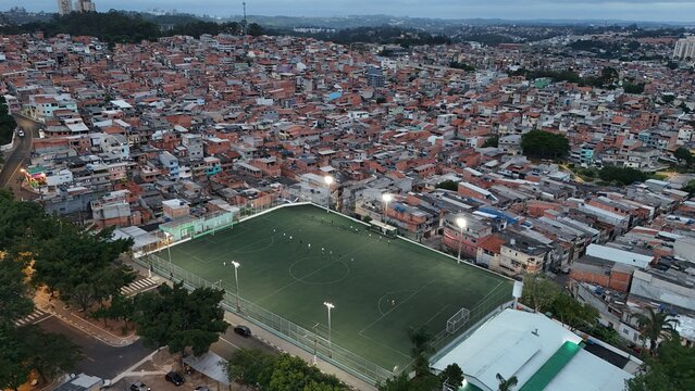 Aerial view of an outdoor soccer field surrounded by dense residential buildings in Osasco, S&atilde;o Paulo, Brazil, with players on the field during the evening
