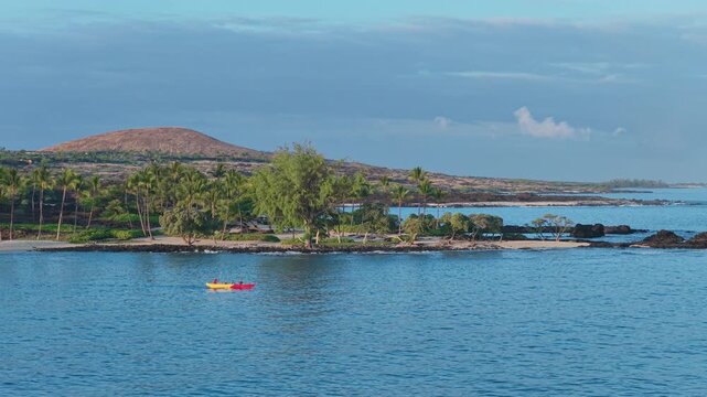 Aerial drone view of two kayakers paddling calm blue ocean water near palm tree lined lava rock shoreline of Big Island Hawaii with volcanic cinder cone hill glowing in morning light.
