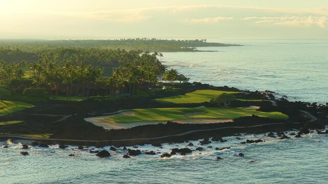 Drone flight over lush green oceanfront golf course on Big Island Hawaii with black lava rock coastline palm trees and turquoise Pacific waves glowing in warm golden sunrise light.