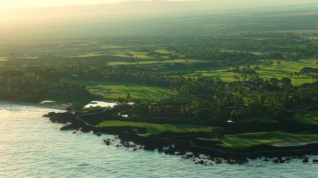 Aerial drone view of luxury coastal golf resort on Big Island Hawaii at golden sunset with palm trees green fairways lava rock shoreline ponds and calm turquoise Pacific ocean waves below.