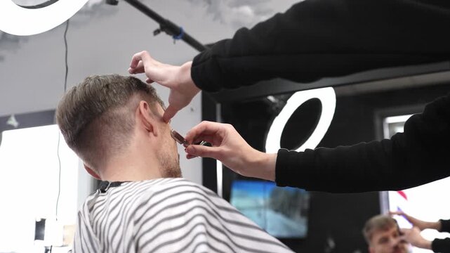 Skilled barber performing precise hair fade using straight razor, working on client's hair in contemporary grooming salon with professional tools