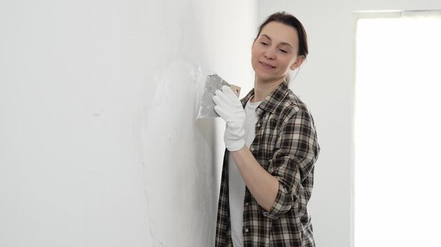 Woman wearing protective gloves and checkered shirt smoothing putty on white wall, spreading compound with spatula during home renovation, displaying practical home improvement skills