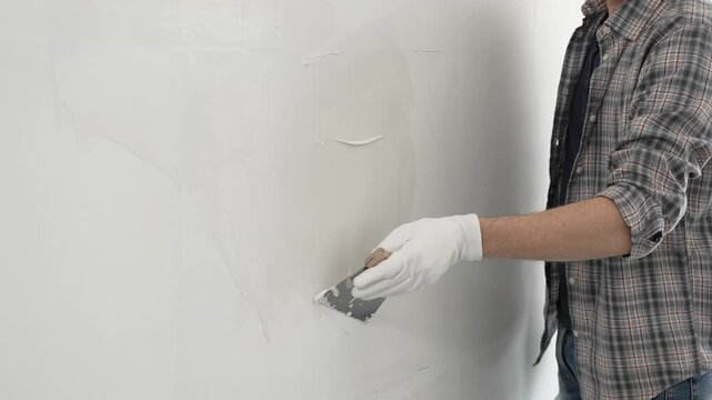 Unknown construction worker man wearing gloves and plaid shirt is using a putty knife to apply plaster on a white wall, during a home renovation or construction project