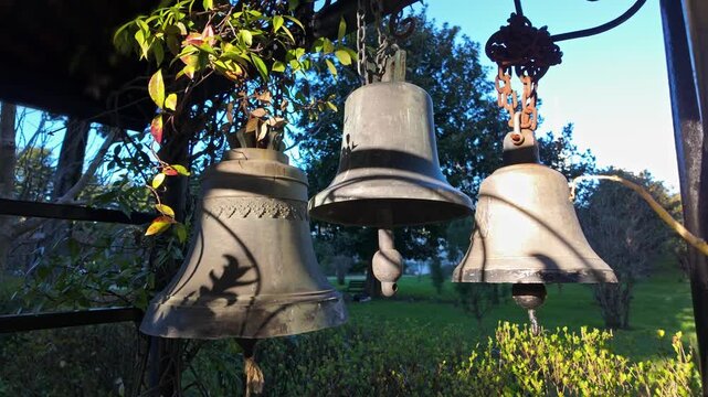 Three old weathered bronze bells hang with rusty chains in a sunny park among green foliage.