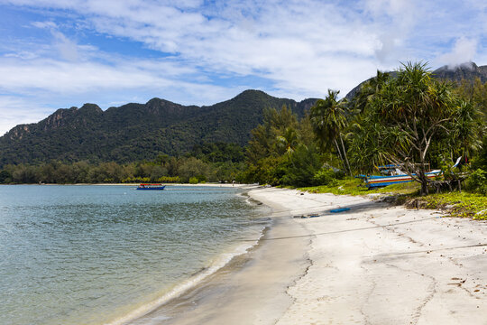 View of Tanjung Rhu Beach with turquoise waters, lush green mountains, palm trees, and a small boat on the sandy shore in Langkawi, Kedah, Malaysia.
