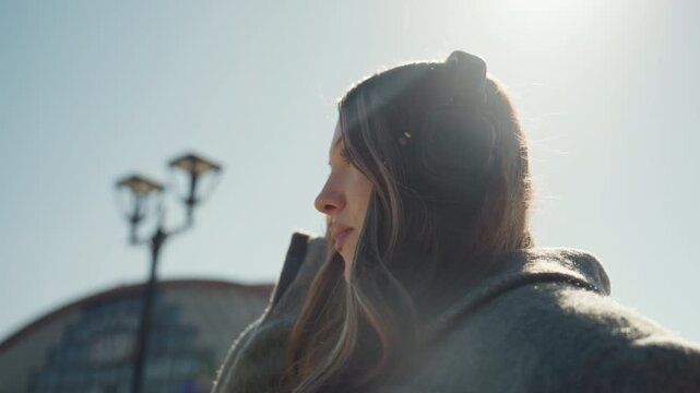 Woman with headphones. Cheerful woman in cozy coat smiling. Candid shot of woman adjusting headphones outdoors. Playful woman with rosy cheeks and headphones under sunlit station backdrop
