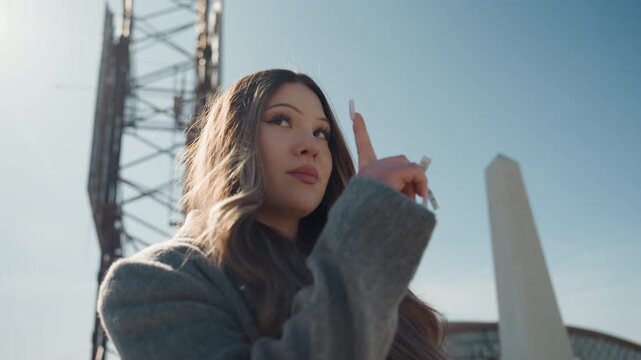 Playful rooftop gesture by asian woman, pretty guide pointing upward with thoughtful smile and warm backlight rim, industrial lattice tower and obelisk framing scene, cozy coat and long hair, hopeful