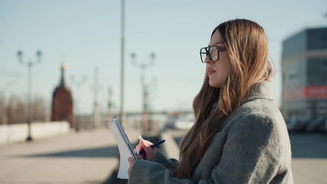 Person reviewing schedule outside. Woman arranging her day plan beneath morning sky outdoors. Thoughtful woman making notes and adjusting glasses in outdoor setting near churches and street lamps