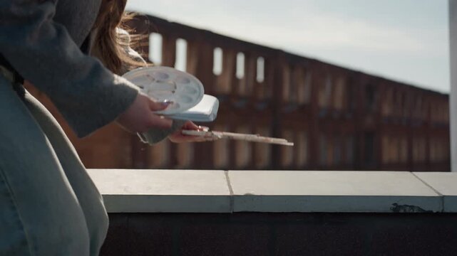 Pretty rooftop sketcher white woman places palette on ledge, arranges watercolor pans and prepares to sit, leans to study brick skyline, cinematic afternoon light and calm reflective posture.