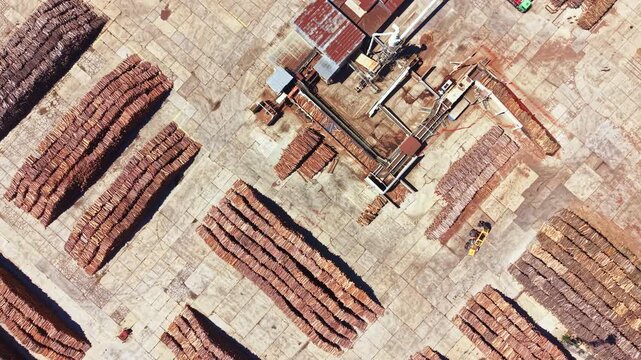 Logs are stacked in neat rows at a wood processing site. Machinery is visible in the center. The area is organized and busy with wood preparation activities.