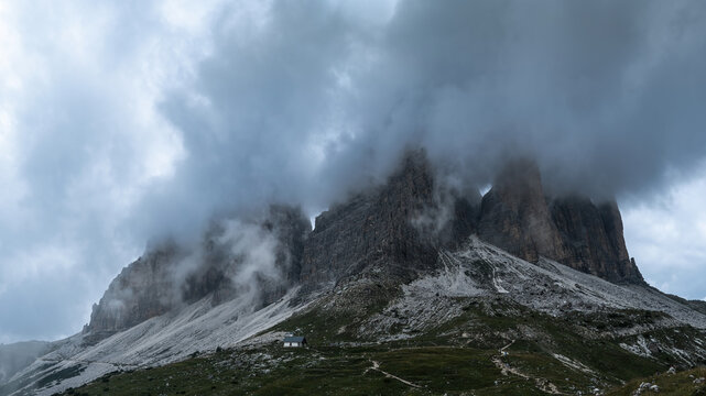 View of Tre Cime di Lavaredo shrouded in thick clouds with rocky peaks and a small building at the base in Italy.