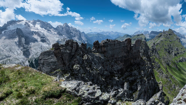 View of Passo Fedaia in the Dolomites, featuring rugged rocky peaks, green alpine meadows, and a clear blue sky with scattered clouds in Rocca Pietore, Veneto, Italy.