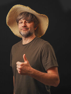 Man in his 40s or 50s wearing green shirt and big straw hat, dark background. Country looking male model with grey beard, glasses and friendly face. Thumbs up positive gesture.