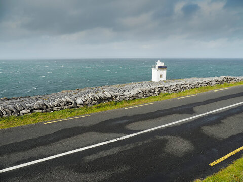 White Black head lighthouse stands on rough stone coast. Burren, Ireland. Ocean stretches wide, meeting sky with clouds. Rocky terrain shows nature raw, untamed strength. Tourist attraction.