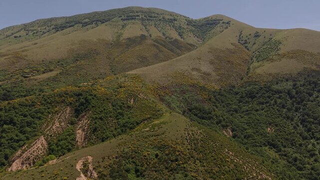 Aerial view of rolling green hills and mountains with patches of yellow vegetation under a clear blue sky in Muzine, Qarku i Vlores, Albania.