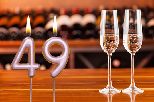 Birthday card, anniversary or celebration of a special day, lit candles forming the number 49 and two champagne glasses; Photo on a background of a bookshelf with blurred wine bottles.