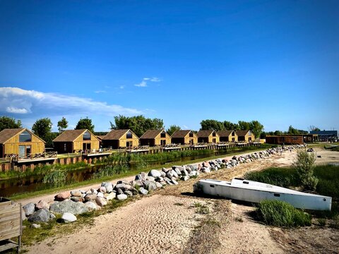 A row of wooden cabins line a tranquil waterway in Dreverna, Klaipeda District, Lithuania. The cabins feature pitched roofs and large windows, set along a constructed pier. 