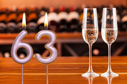 Birthday card, anniversary or celebration of a special day, lit candles forming the number 63 and two champagne glasses; Photo on a background of a bookshelf with blurred wine bottles.