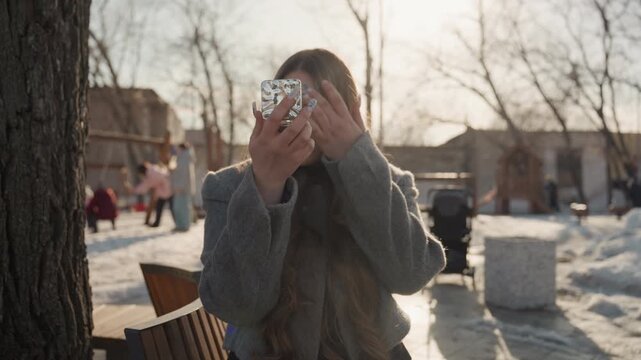 Elegant Asian woman shyly covering face. Charming Asian female in winter attire relaxing on park bench. Graceful Asian woman with gentle smile adjusting coat outdoors in park setting