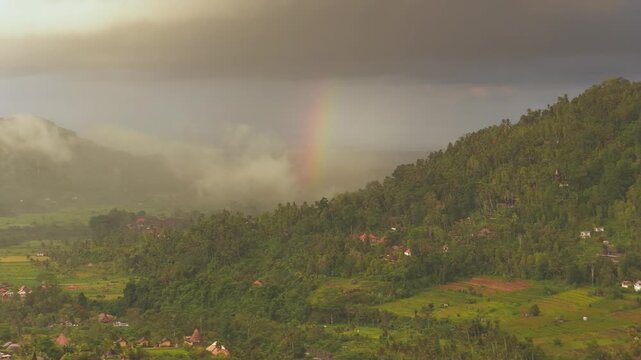 Aerial view of Sidemen Rice Terrace with a rainbow over lush green hills and tropical vegetation in Kecamatan Sidemen, Bali, Indonesia.