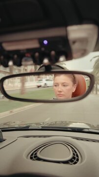 Focused woman driving a modern convertible car during a sunny day trip, her face visible in the rearview mirror as she navigates the road with a calm and concentrated expression. Vertical video