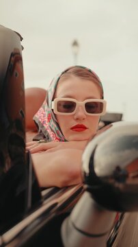 Beautiful young woman with red lips wearing a headscarf and sunglasses leaning on the door of a classic black convertible, embodying a sense of vintage style, freedom, and luxury travel