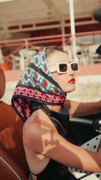 Beautiful woman wearing a headscarf and sunglasses driving a classic convertible car during a sunny day, embodying a concept of vintage elegance, freedom, and luxurious travel. Vertical video