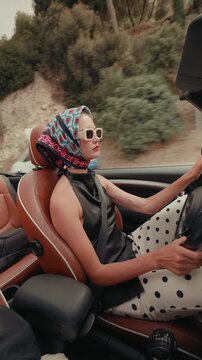 Fashionable young woman wearing a headscarf and sunglasses driving a modern convertible car on a scenic road trip, enjoying the freedom and luxury of her summer vacation adventure. Vertical video
