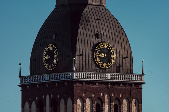 View of the historic clock tower of Riga Cathedral with its dark metal dome and Roman numeral clock faces against a clear blue sky in Riga, Latvia.