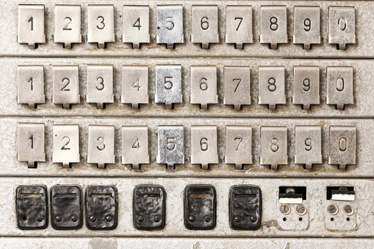 Old numbered buttons cover metal panel. Mechanical keys display worn digits. Dark switches line lower section. Worn controls suggest vintage machinery. Industrial interface forms retro background.